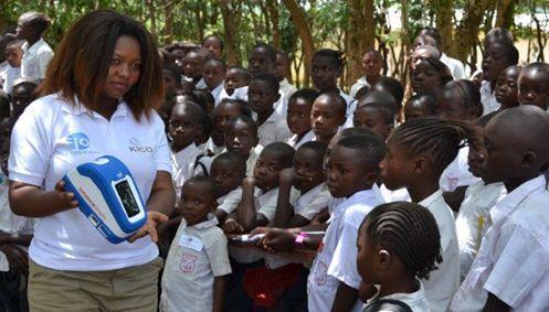 A representative of the Know for Sure initiative, holding a Deki Reader used to conduct rapid diagnostic testing for malaria, explains to children how to get access to testing for themselves and their parents.